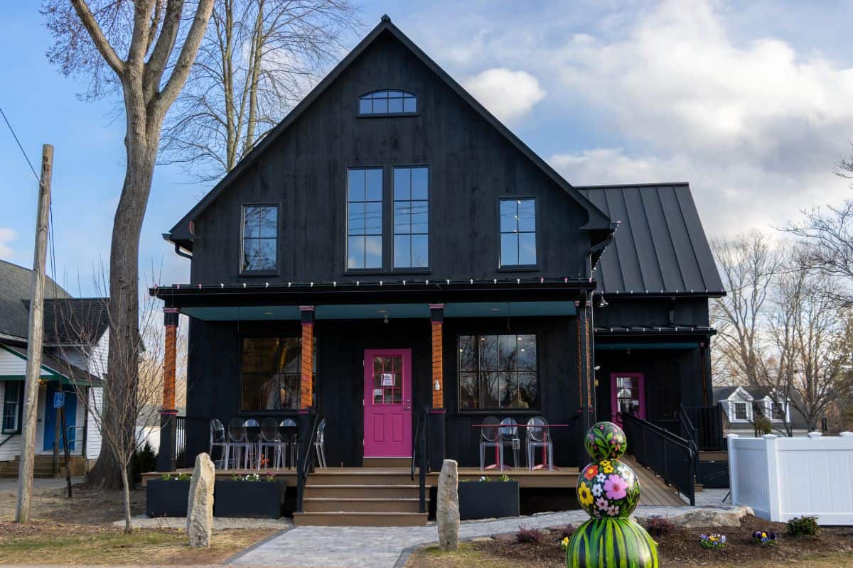 A black house with a dark metal roof, large windows, and a bright pink front door, featuring a front porch and outdoor seating.