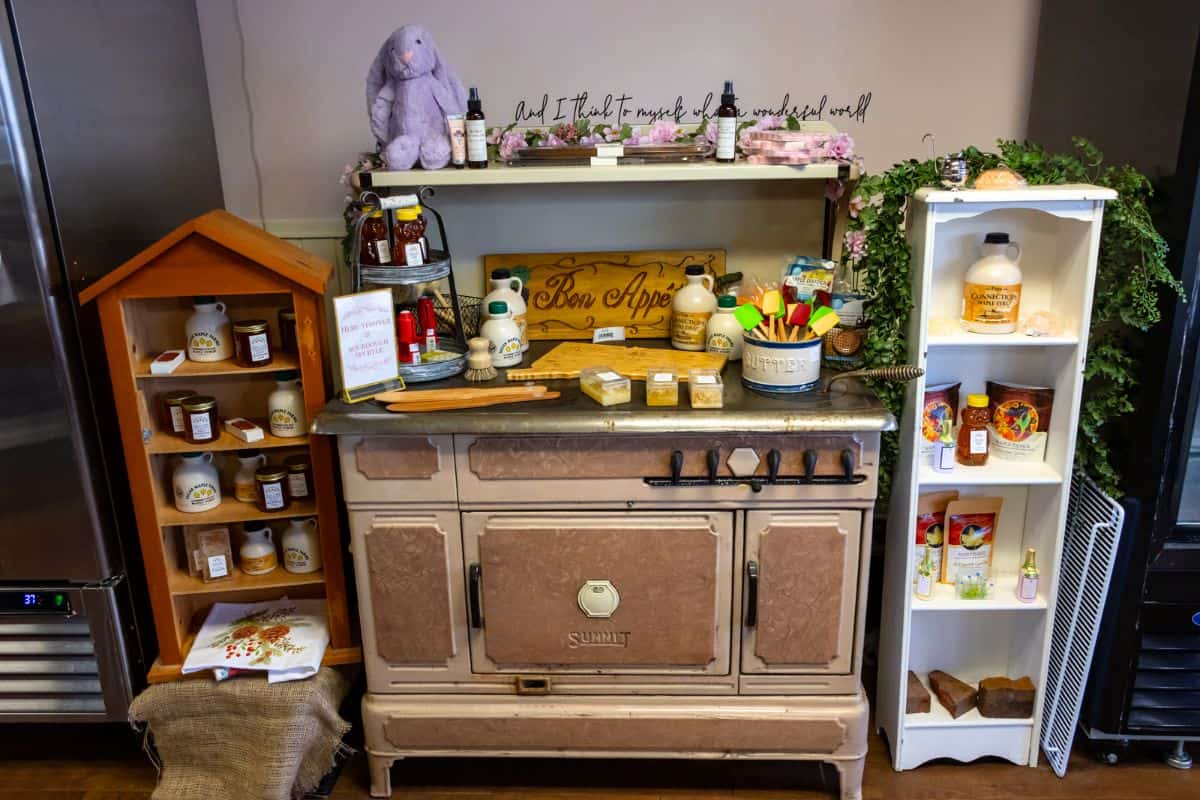A vintage stovetop display holds kitchen utensils, honey jars, sauces, a plush toy, and decorative items, surrounded by small shelves with more food products and greenery.