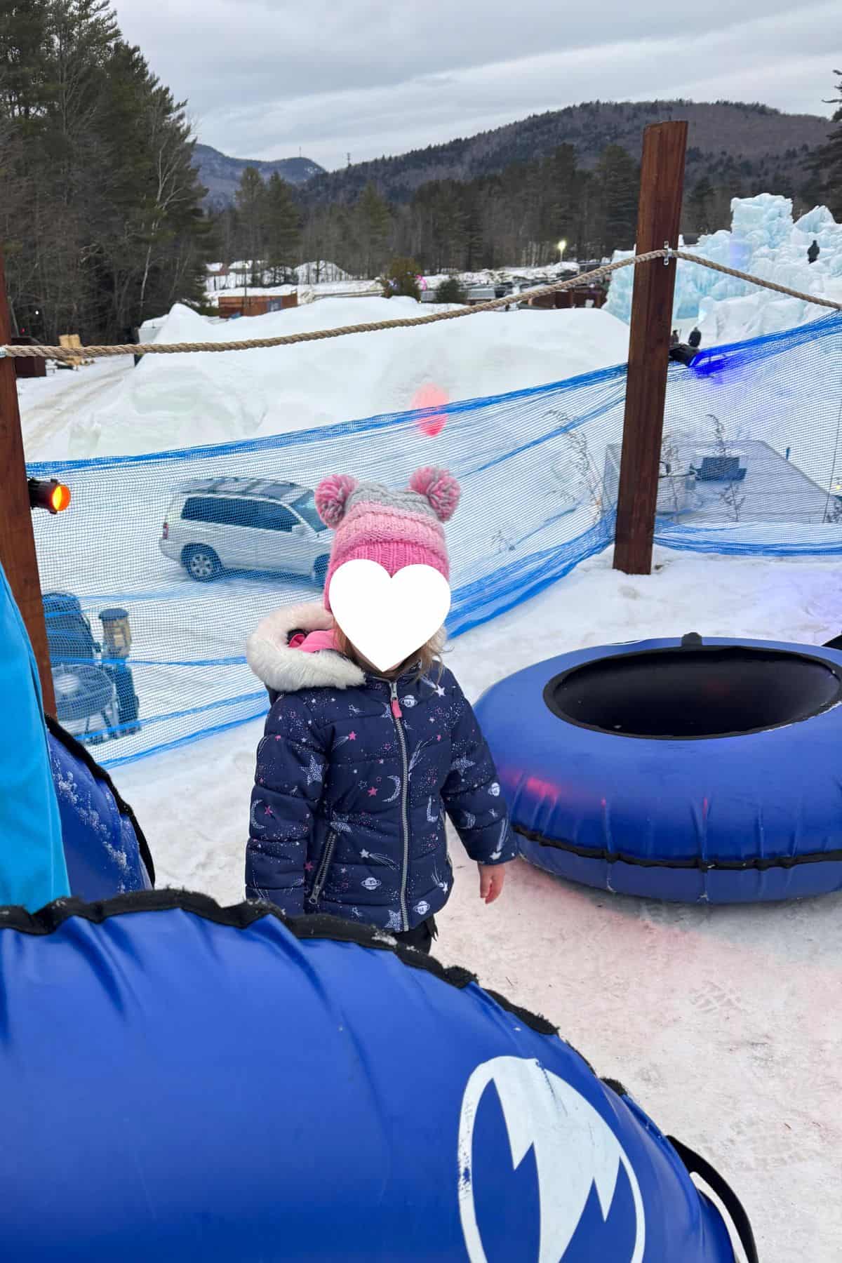 A child in a winter coat stands on snow near blue snow tubes, with an icy landscape, fencing, and parked cars in the background.
