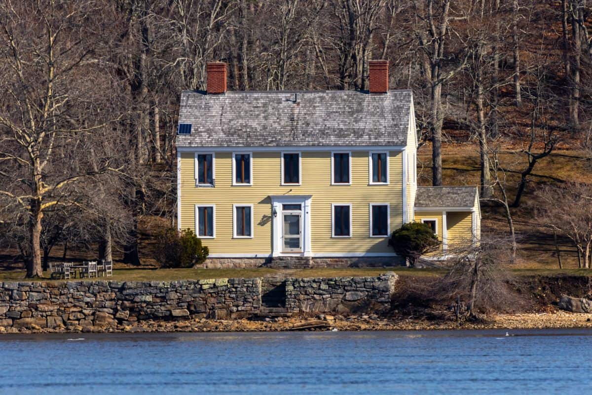 A yellow two-story house with a stone foundation stands near the water, surrounded by leafless trees and a grassy area.