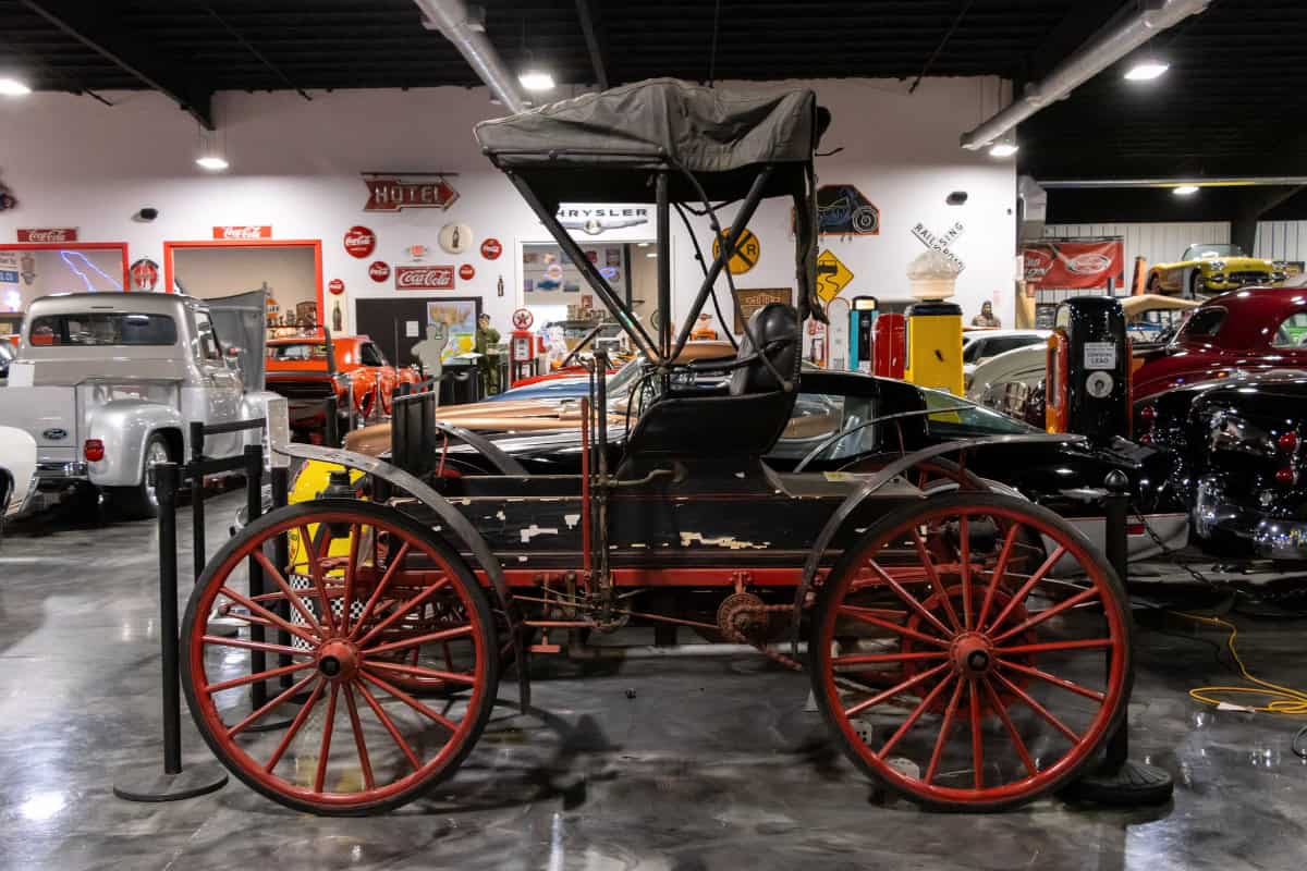 An antique black and red horse-drawn carriage is displayed indoors among classic cars and vintage signs in a museum setting.