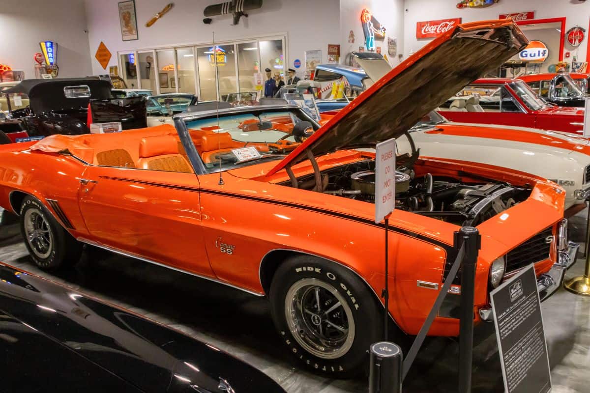 An orange vintage Chevrolet Camaro SS convertible with its hood open is displayed indoors among other classic cars.
