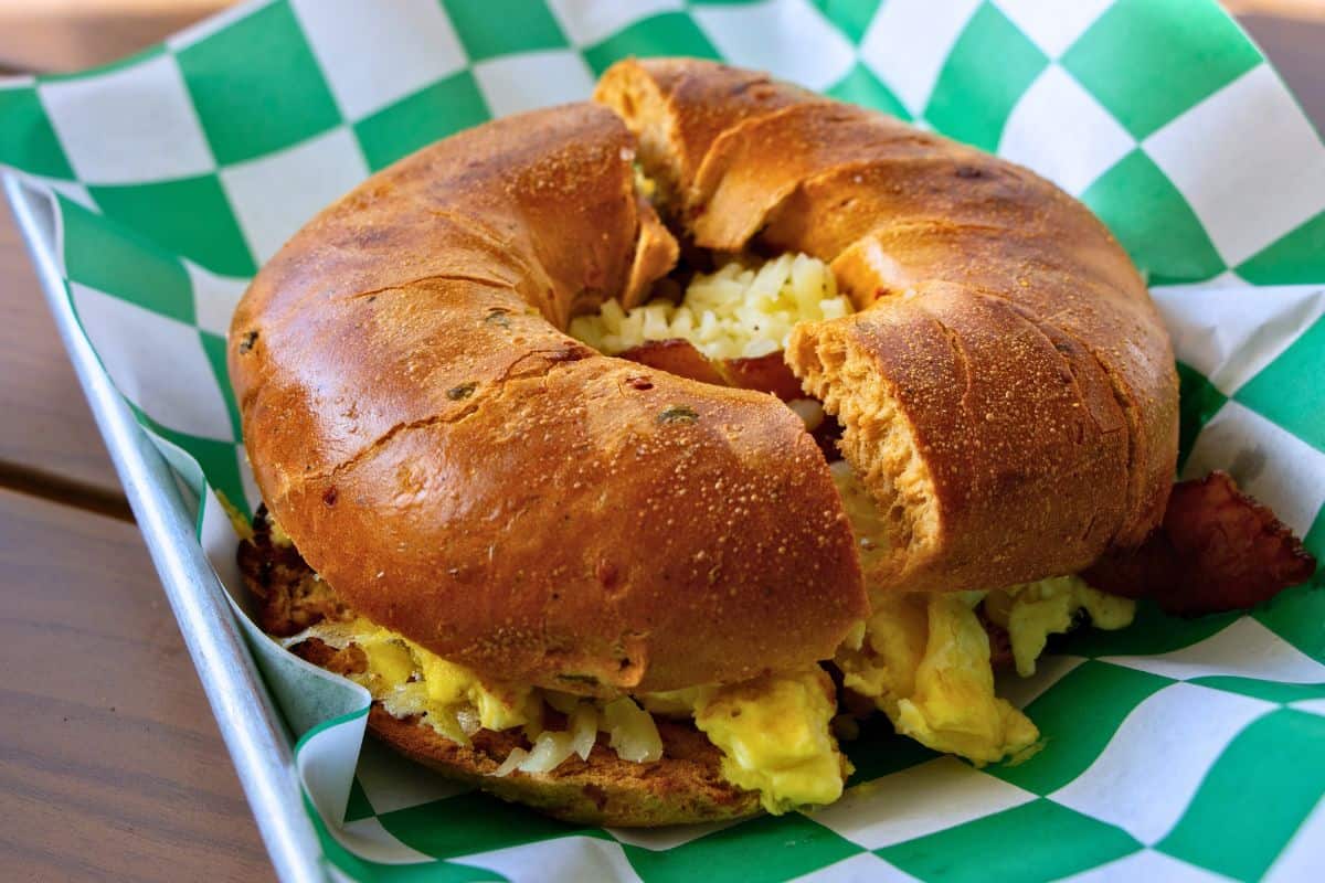 A bagel sandwich filled with scrambled eggs, shredded cheese, and bacon sits on a green and white checkered paper in a metal tray—perfect fuel before exploring Fort Matanzas National Monument.