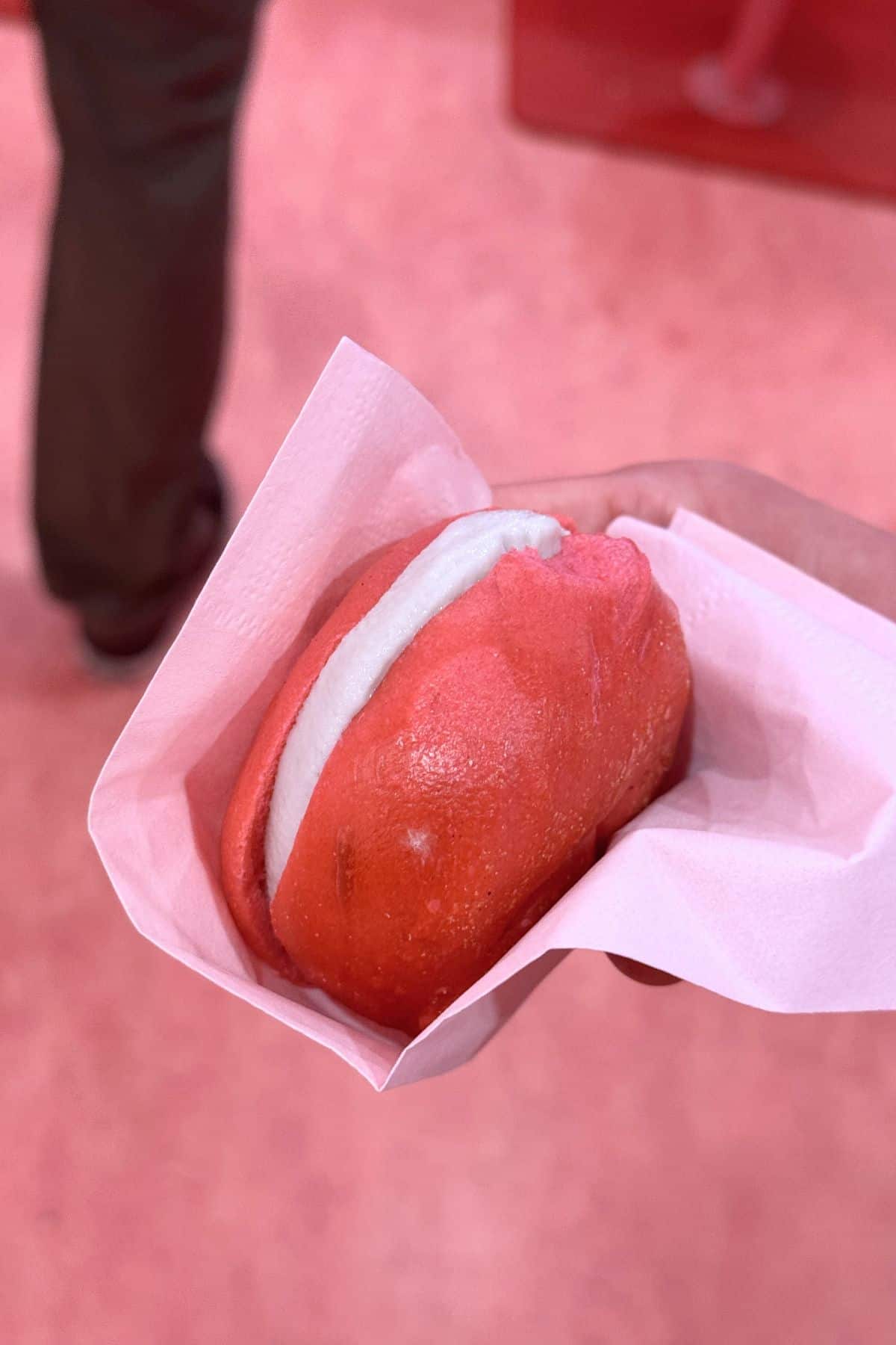 A hand holds a pink steamed bun filled with white cream, wrapped in a pink napkin. A person stands in the background on a pink floor.