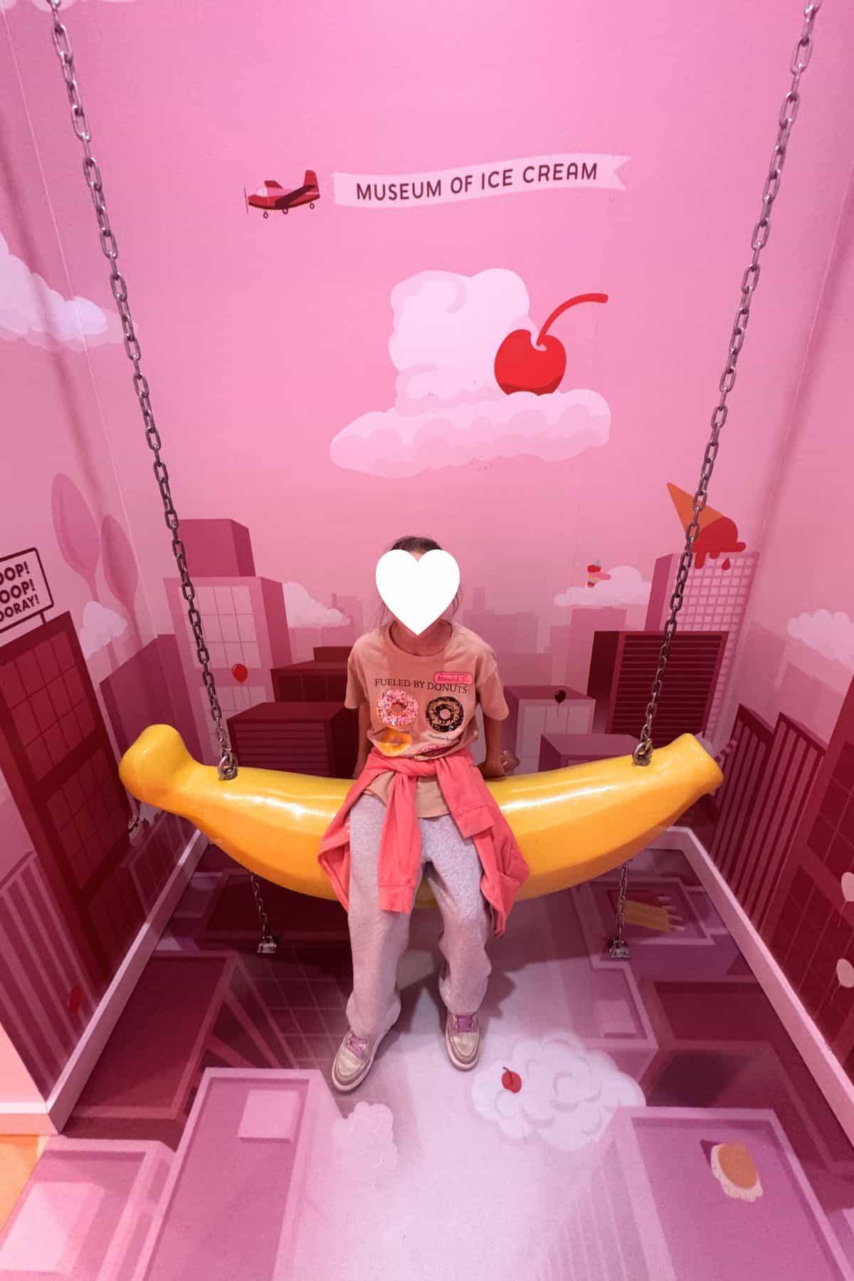 A person sits on a yellow banana-shaped swing in a pink room themed after the Museum of Ice Cream, with cartoon cityscape and cherry illustrations on the walls.
