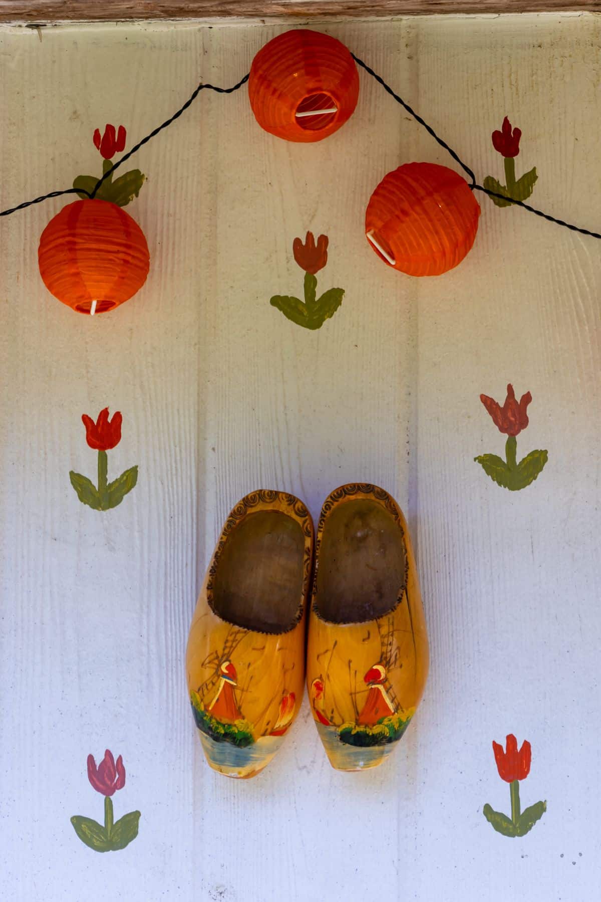 A pair of decorative wooden clogs hangs on a white wall adorned with painted red tulips, beneath three hanging orange paper lanterns.