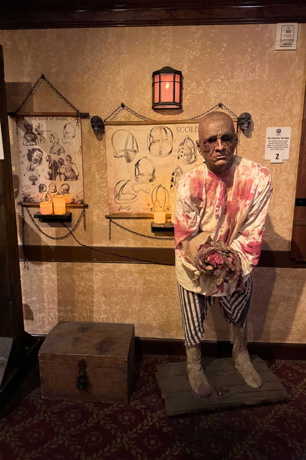 A mannequin dressed as a bloodied prisoner stands in front of historical torture device illustrations and props in a dimly lit museum display.