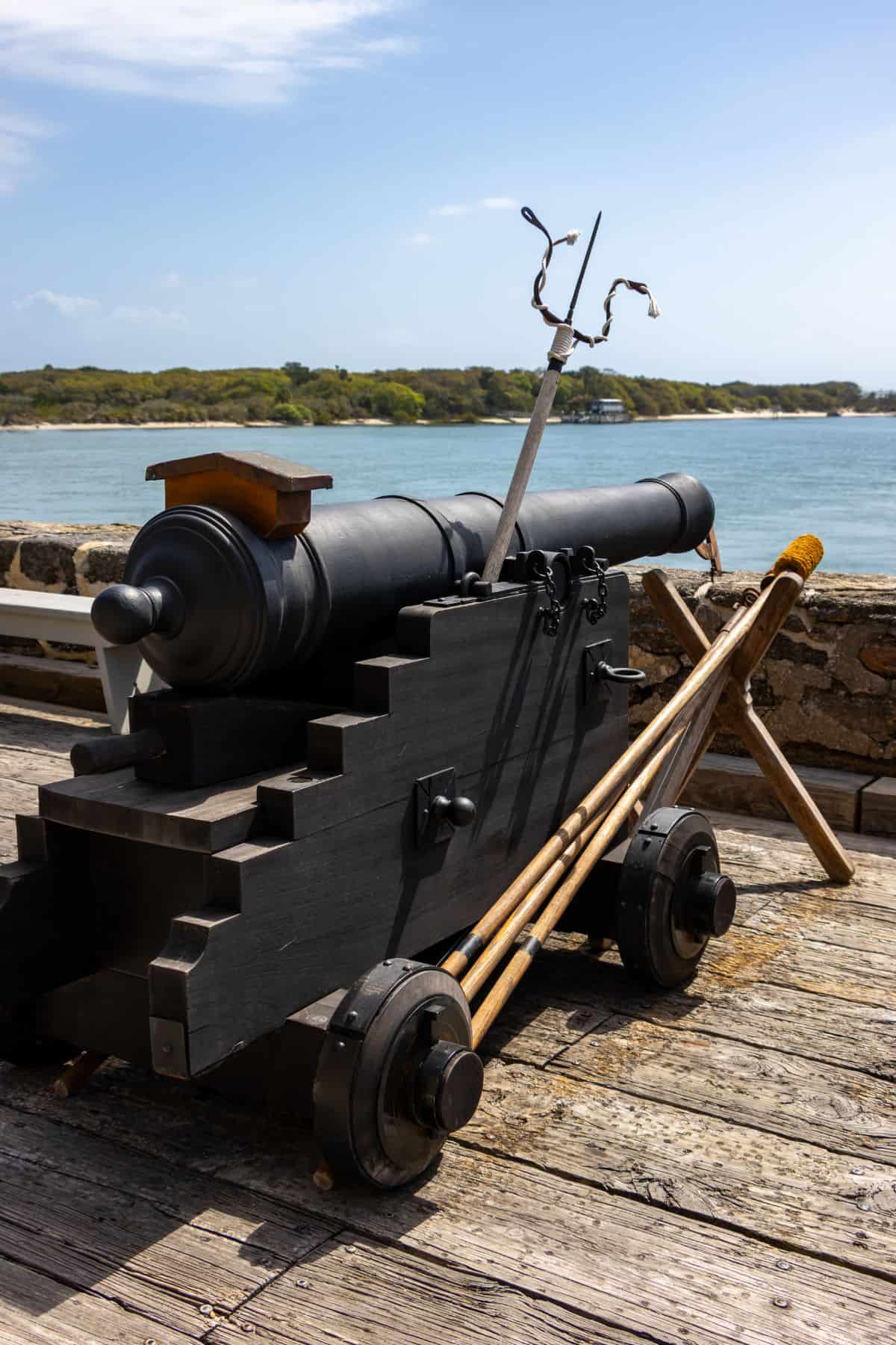 A black cannon on a wooden carriage is positioned on a wooden deck beside water, with cleaning tools and greenery in the background.