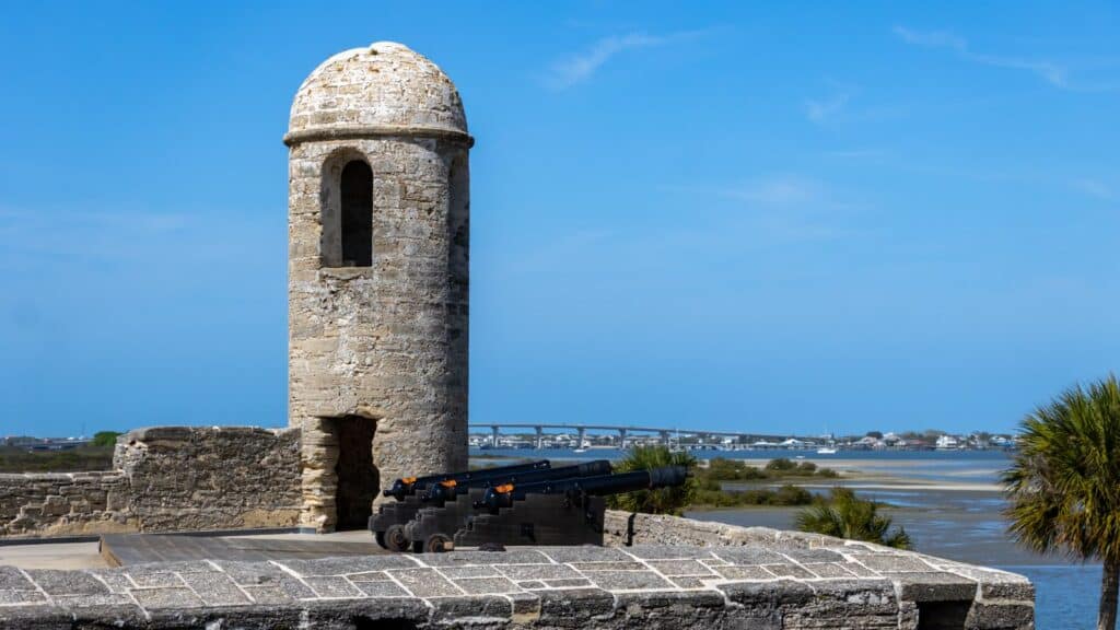 Stone watchtower with a cannon overlooks a river, with a bridge and buildings visible in the background under a clear blue sky.