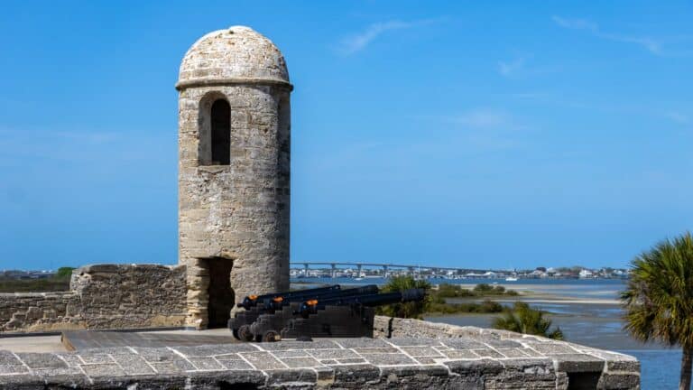 Stone watchtower with a cannon overlooks a river, with a bridge and buildings visible in the background under a clear blue sky.