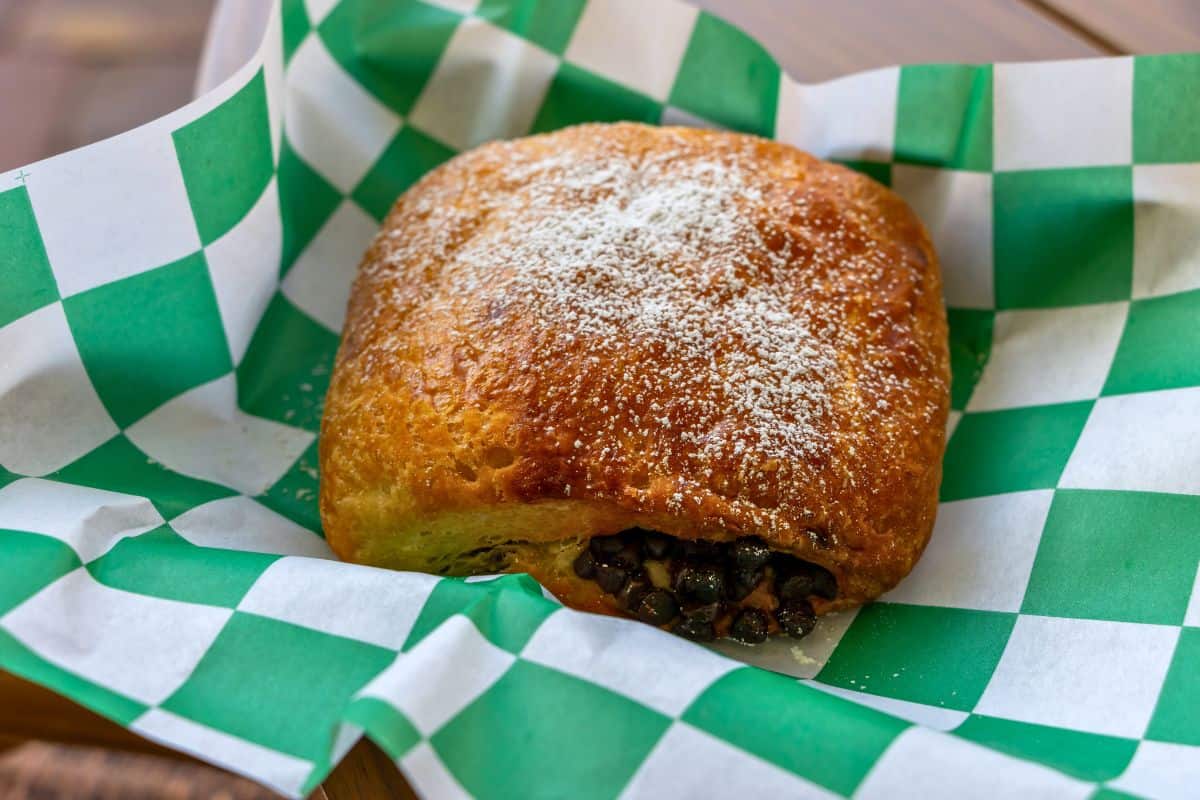 A golden-brown pastry dusted with powdered sugar sits on green and white checkered paper, with a filling visible at one end—perfect for a picnic near Fort Matanzas National Monument.