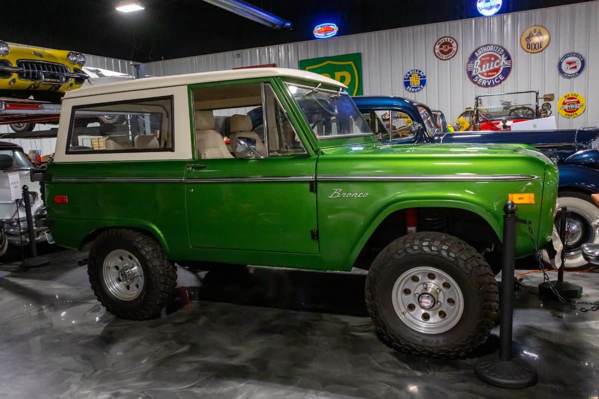 A vintage green Ford Bronco is displayed indoors among other classic cars, with automotive signs on the walls in the background.