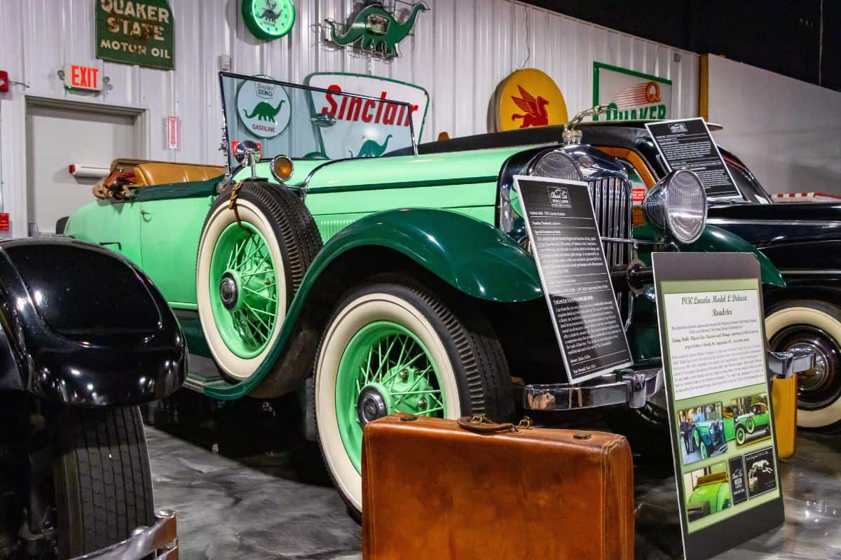 A vintage green and black convertible car is displayed indoors, surrounded by signage and historical information, with a brown suitcase placed in front.
