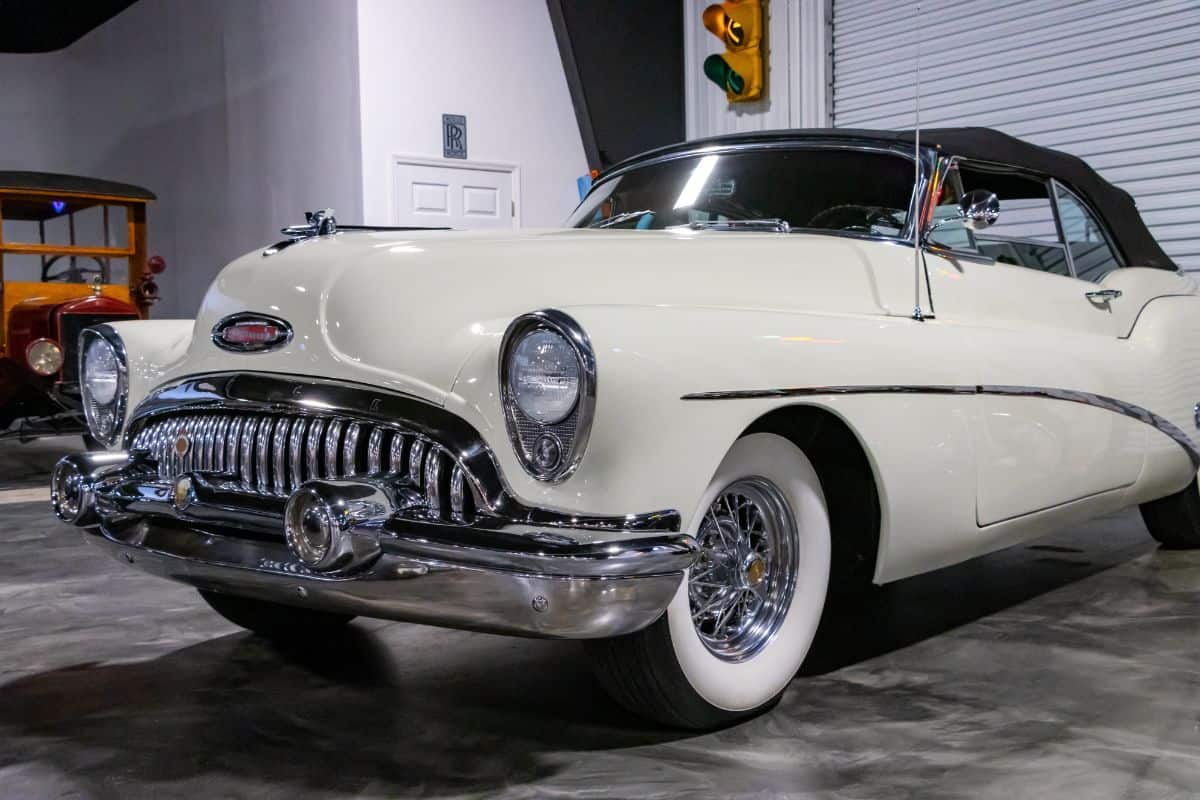 A white vintage convertible car with chrome detailing is parked indoors near a roll-up door and traffic signal.