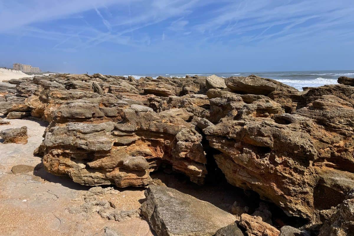 Rocky shoreline with large, weathered brown rocks under a blue sky with wispy clouds and ocean waves in the background at Fort Matanzas National Monument.