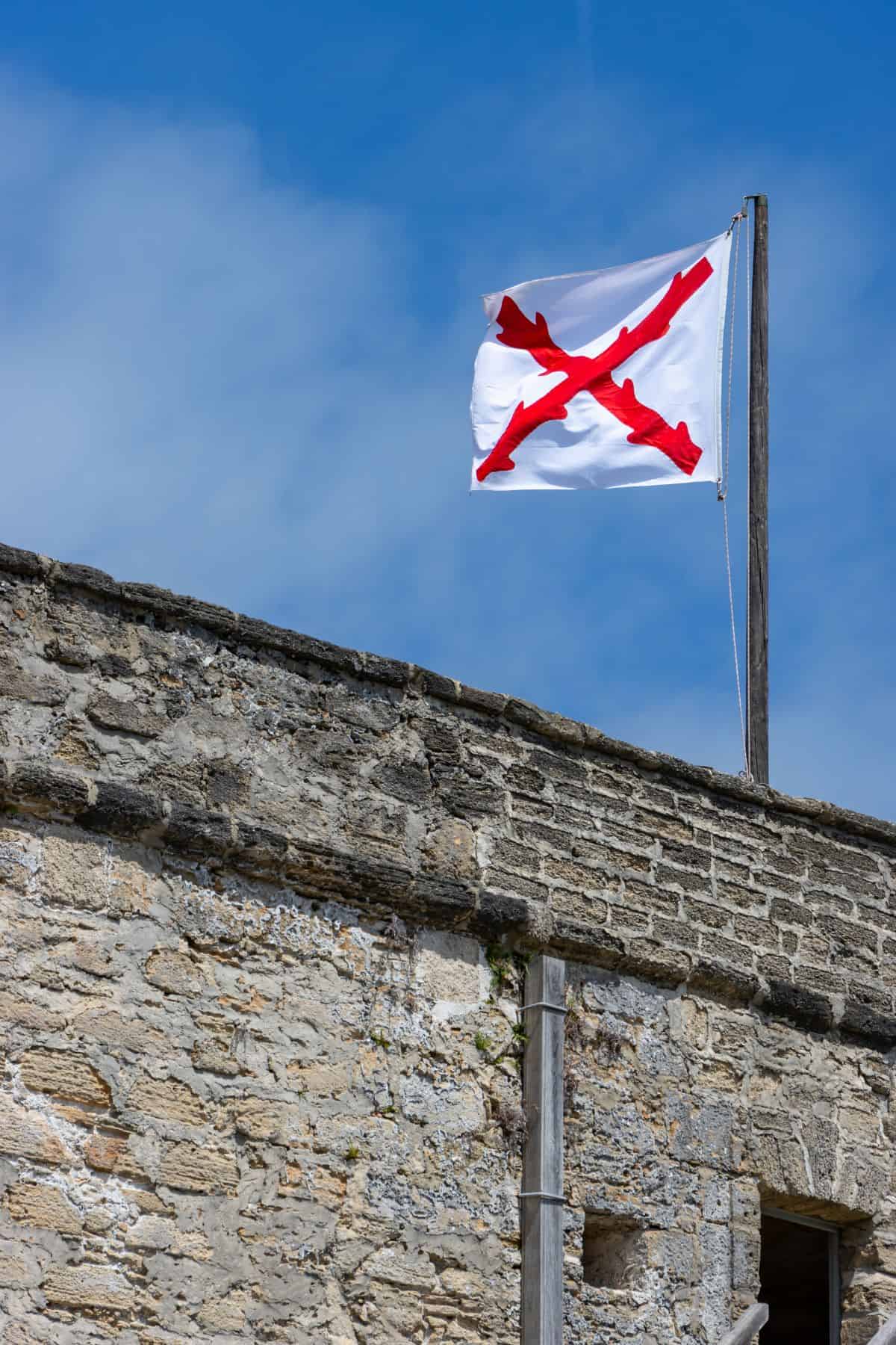 A white flag with a red diagonal cross flies on a pole above a stone building under a blue sky.