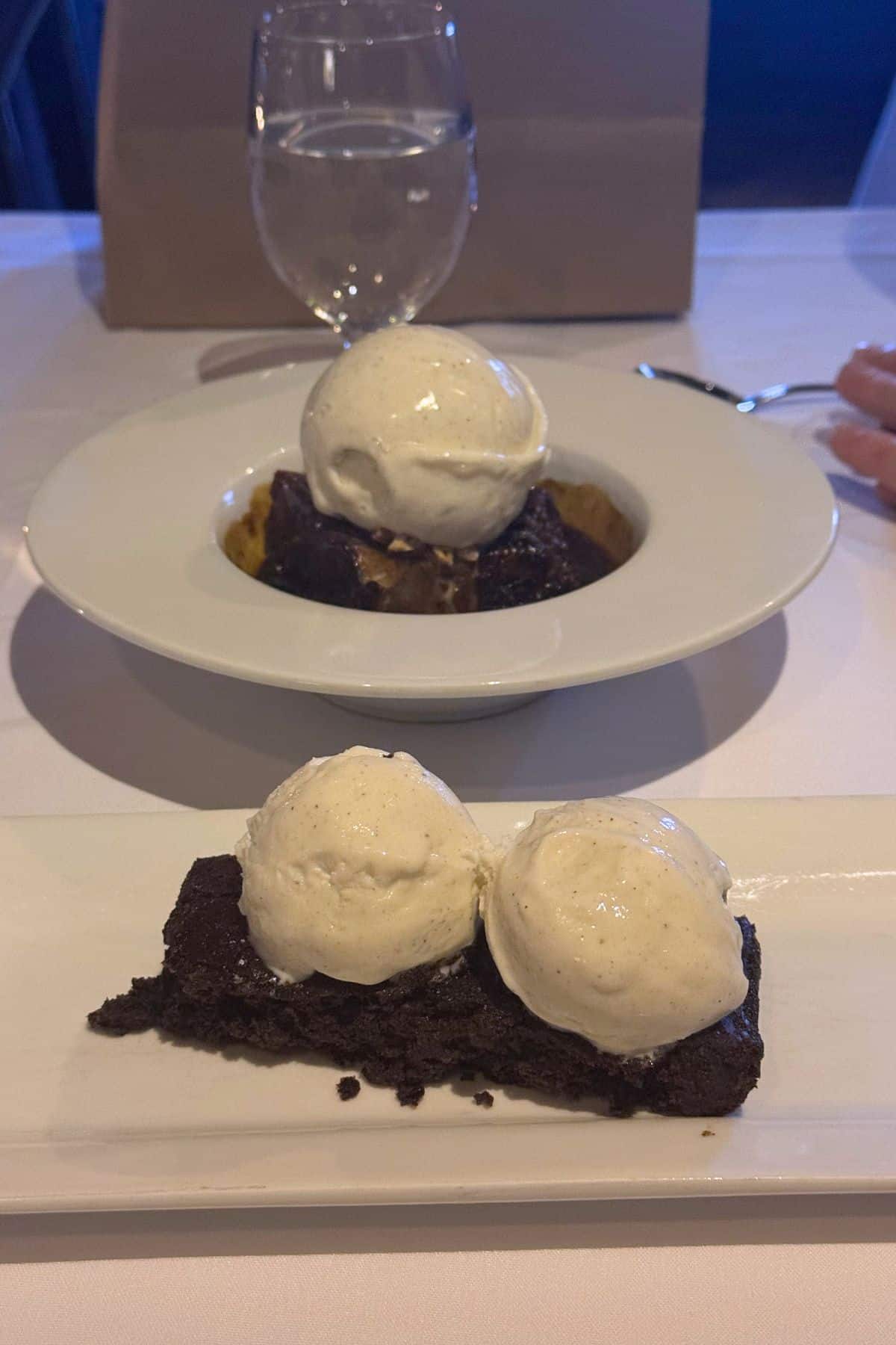A plate with a brownie topped with two scoops of vanilla ice cream in the foreground, and a bowl with a brownie and one scoop of vanilla ice cream in the background.