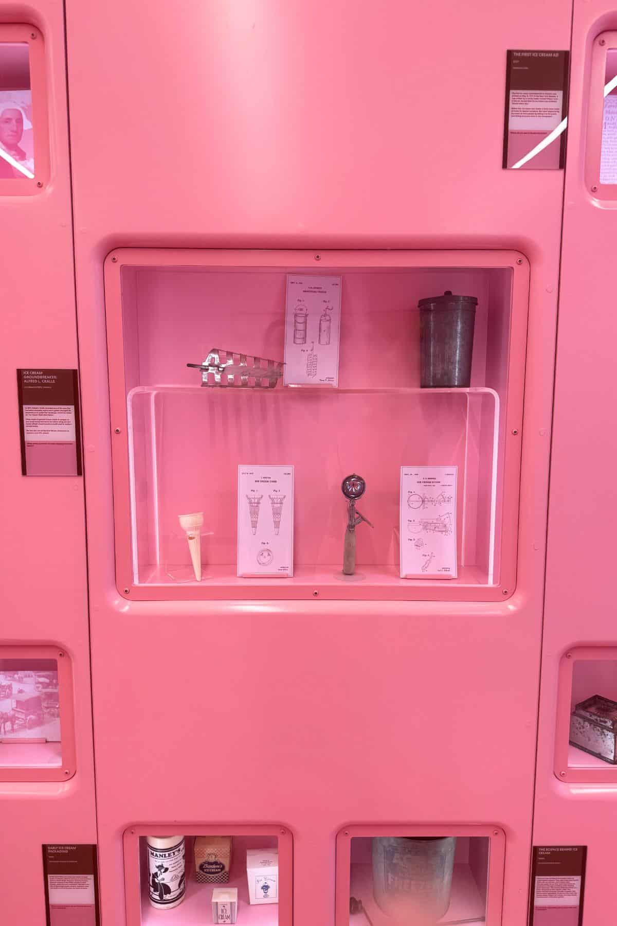 A display case in a pink wall exhibits vintage ice cream scoops, a metal mold, and labeled cards describing each item.