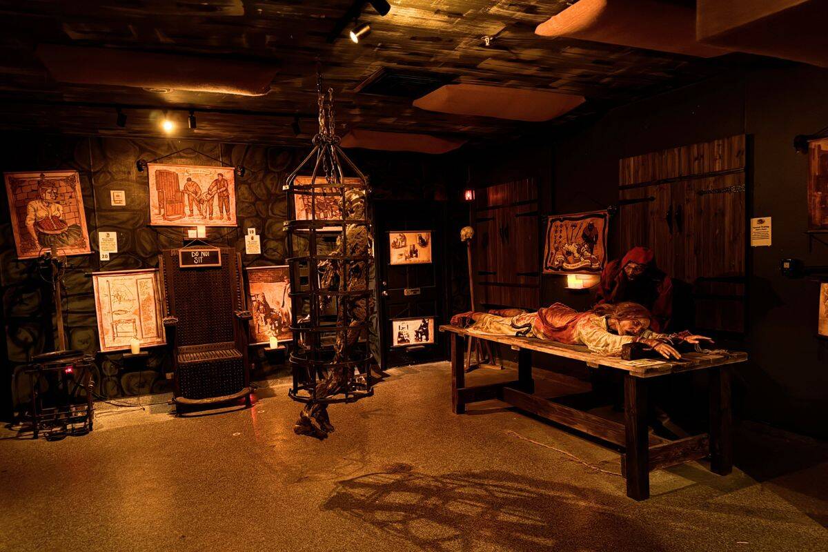 Dimly lit museum exhibit displays medieval torture devices, including an iron cage, torture chair, and a table with a mannequin figure, surrounded by historical illustrations on the walls.