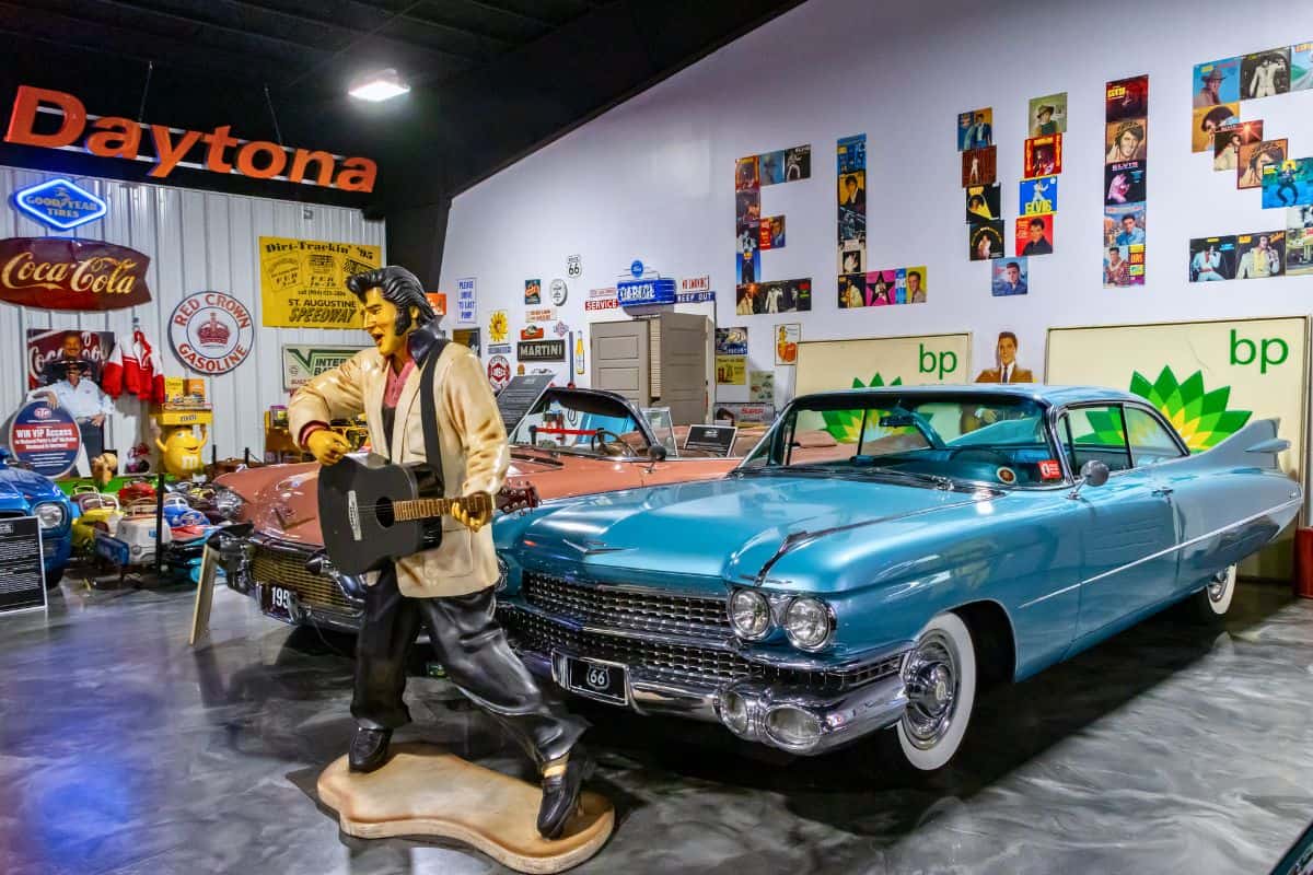 A vintage car museum display with classic cars, a life-size Elvis Presley statue, retro signs, and memorabilia on the walls.