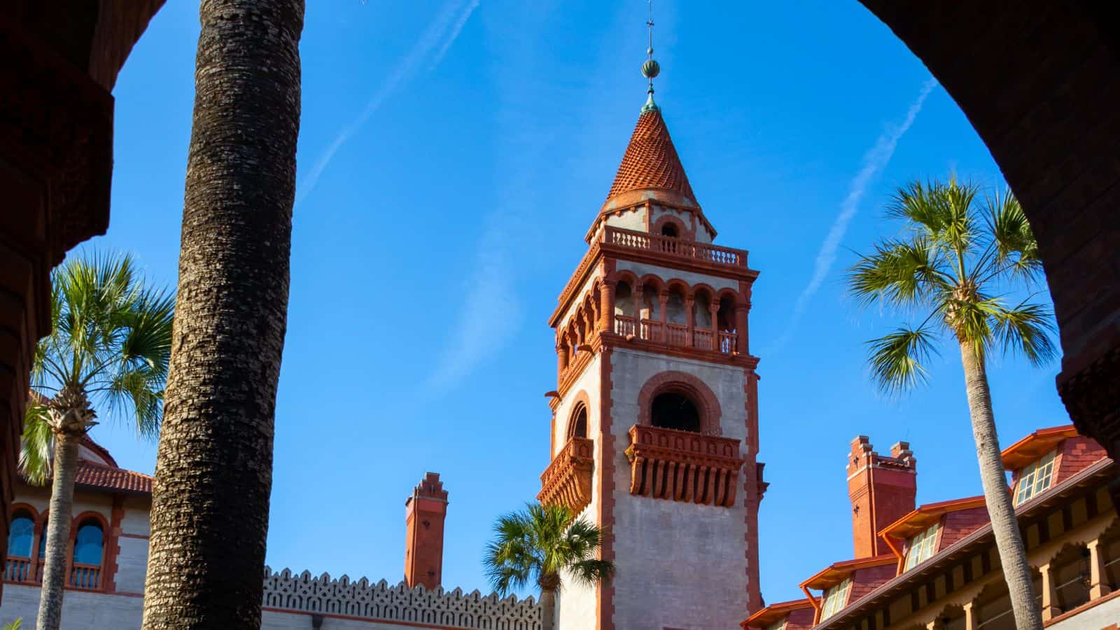A tall, ornate tower with red-tiled roof and arched windows rises among palm trees against a clear blue sky.