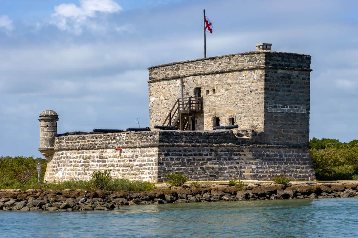 A stone fort with a tower and cannon openings stands by the water, topped with a flag, surrounded by low vegetation and a partly cloudy sky.