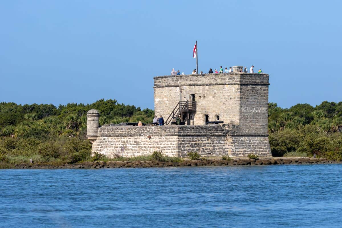 A stone fort with people on top sits by the water, surrounded by greenery, under a clear blue sky.