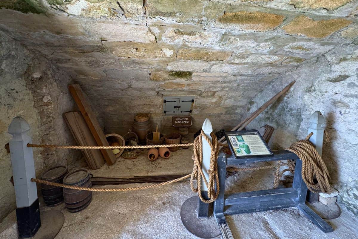Stone-walled storage area with barrels, jars, wooden planks, coiled ropes, and information plaques, sectioned off by ropes and posts.