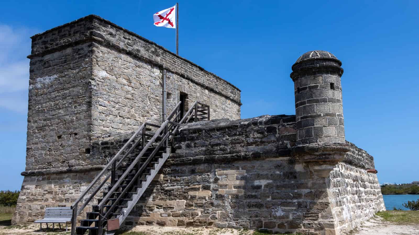 Stone fort with an external wooden staircase, small turret, and a flag with a red cross on top, set against a blue sky.