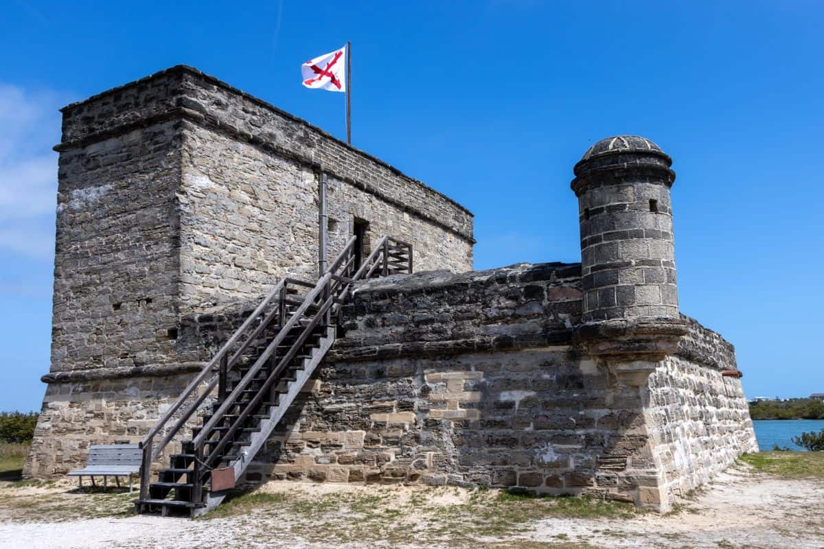 A historic stone fort with a wooden staircase and a white flag featuring a red X, set against a blue sky.