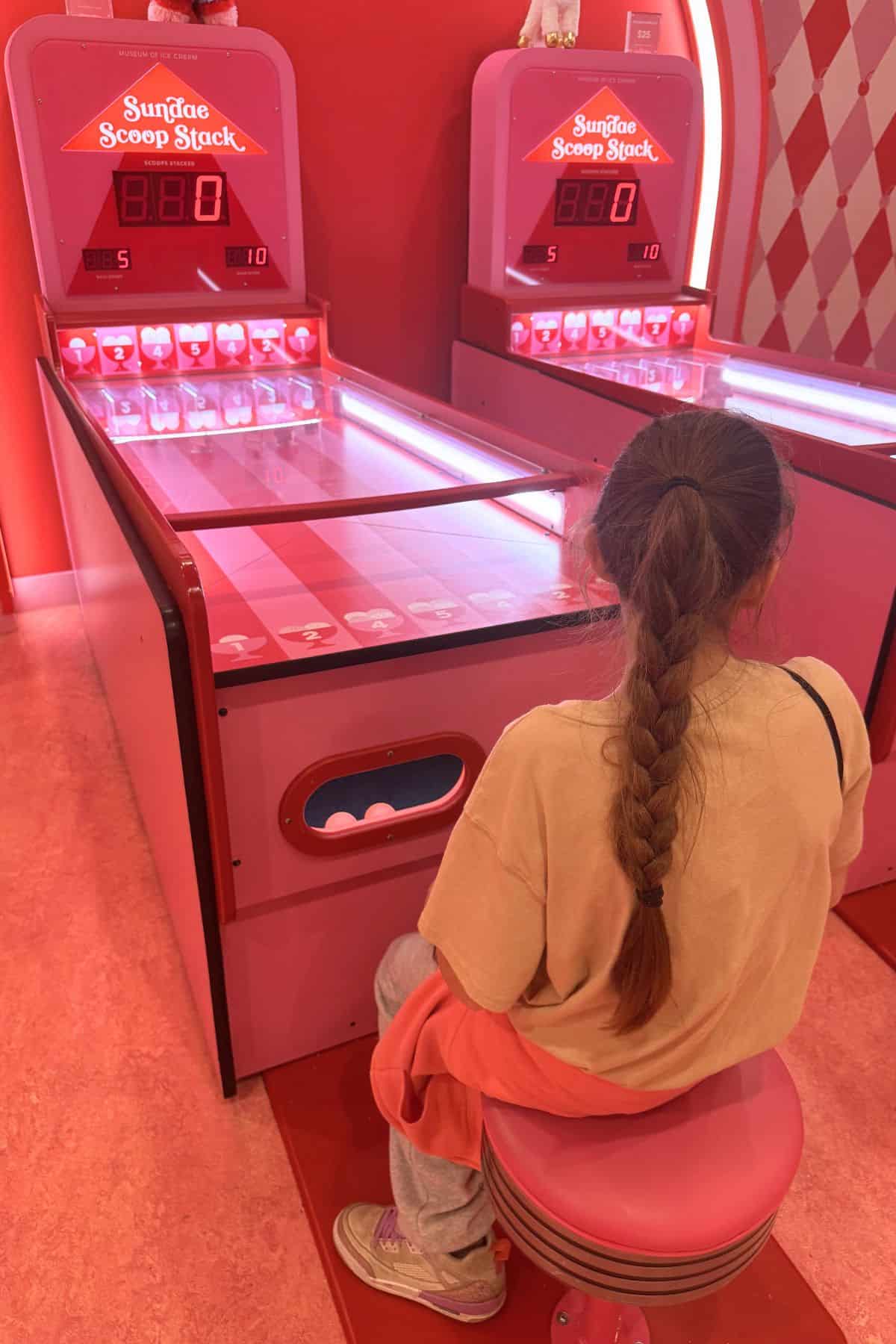 A person with a long braid sits on a pink stool facing a pink arcade skee-ball machine in a brightly lit game room.