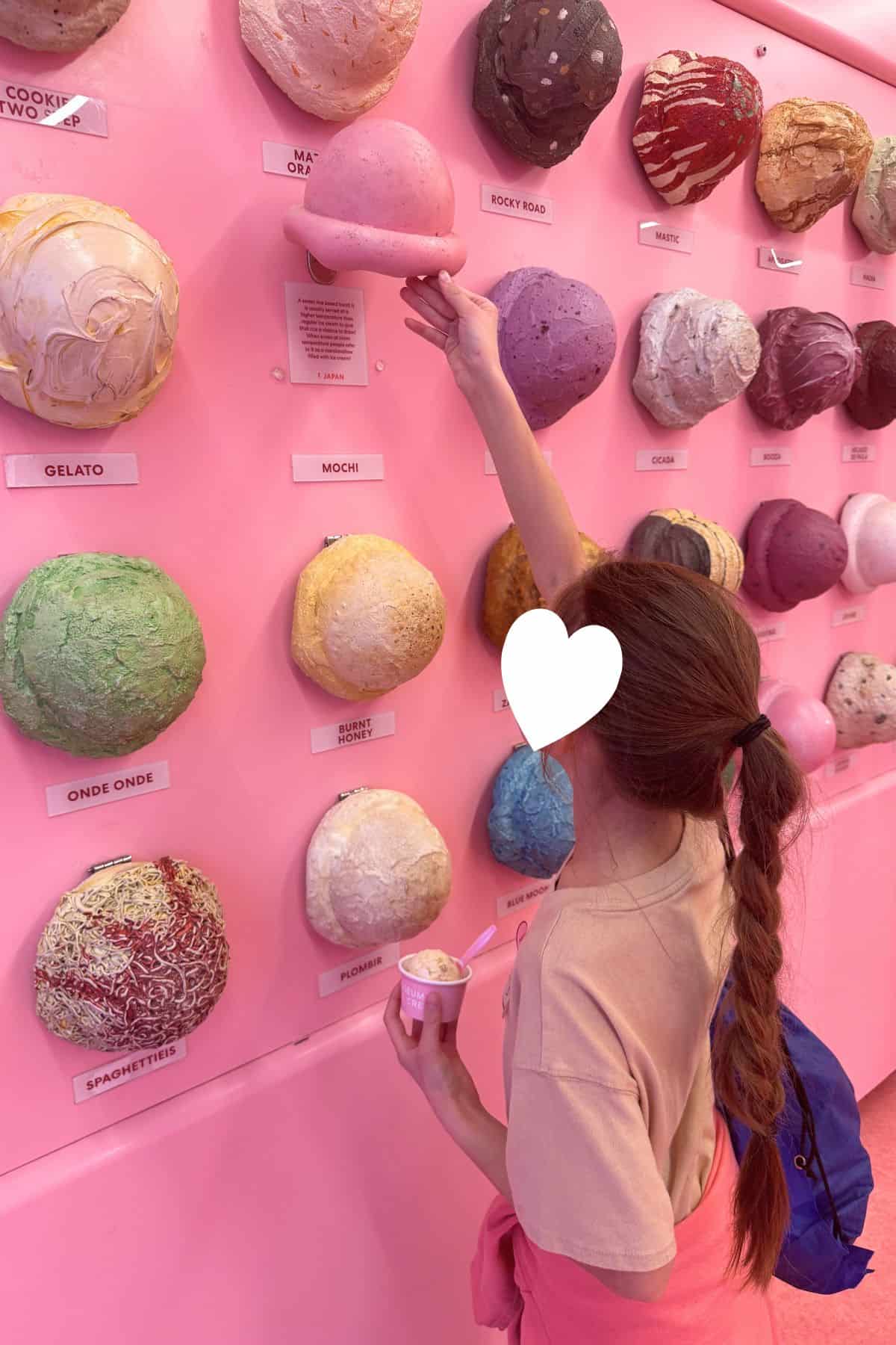 A girl with a long braid and a blue backpack selects a pink mochi sample from a display of ice cream models on a pink wall, holding a cup of ice cream in her other hand.