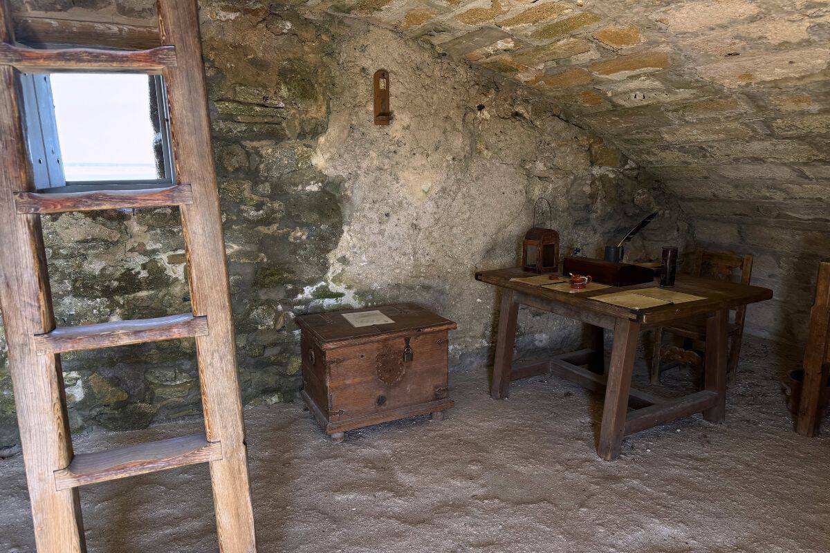 A rustic stone room with a wooden ladder, a chest, and a wooden table holding papers, a candle, and an inkwell. The floor appears to be bare earth.