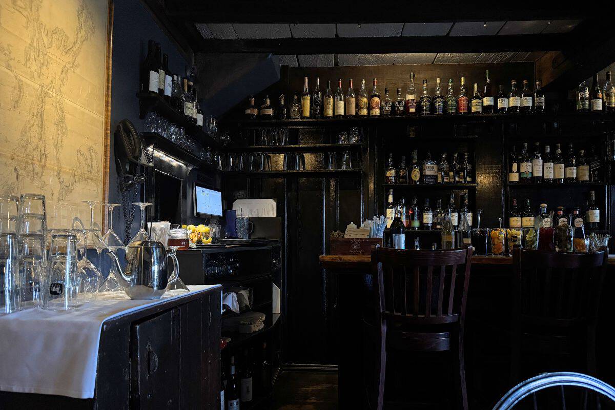Dimly lit bar with dark wooden shelves filled with liquor bottles, a counter with bar stools, and a small side table holding glassware and a metal pitcher.