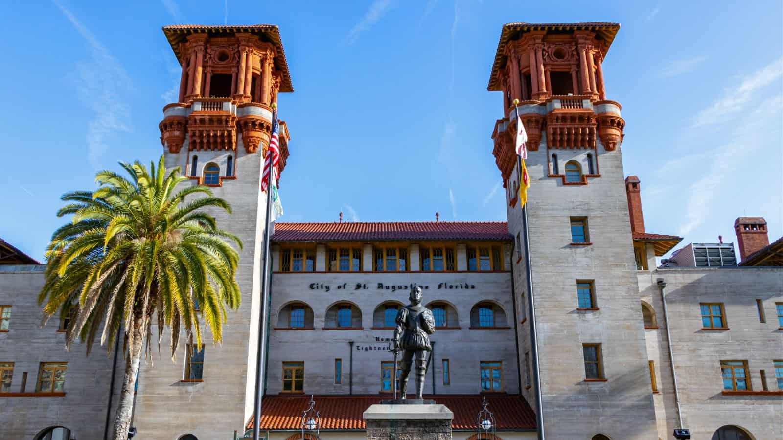 Historic building with two towers, a palm tree on the left, American flags, and a statue in front. The building reads "City of St. Augustine, Florida" above the entrance.