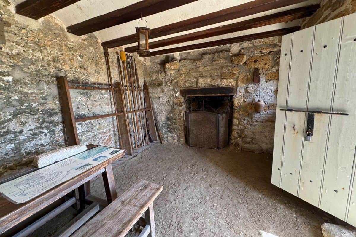 Room with stone walls, wooden ceiling beams, an old fireplace, wooden benches and table with papers, barred window, and an open wooden door.