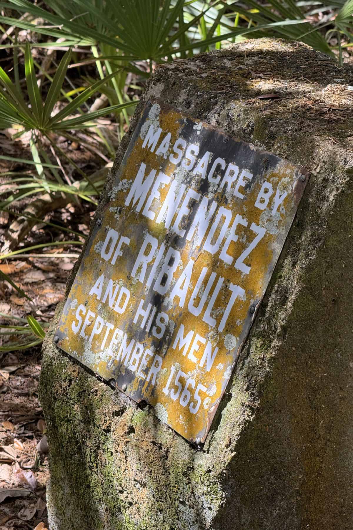 Weathered metal sign on a rock at Fort Matanzas National Monument reads: "Massacre by Menendez of Ribault and his men, September 1565." Surrounded by foliage and sunlight.