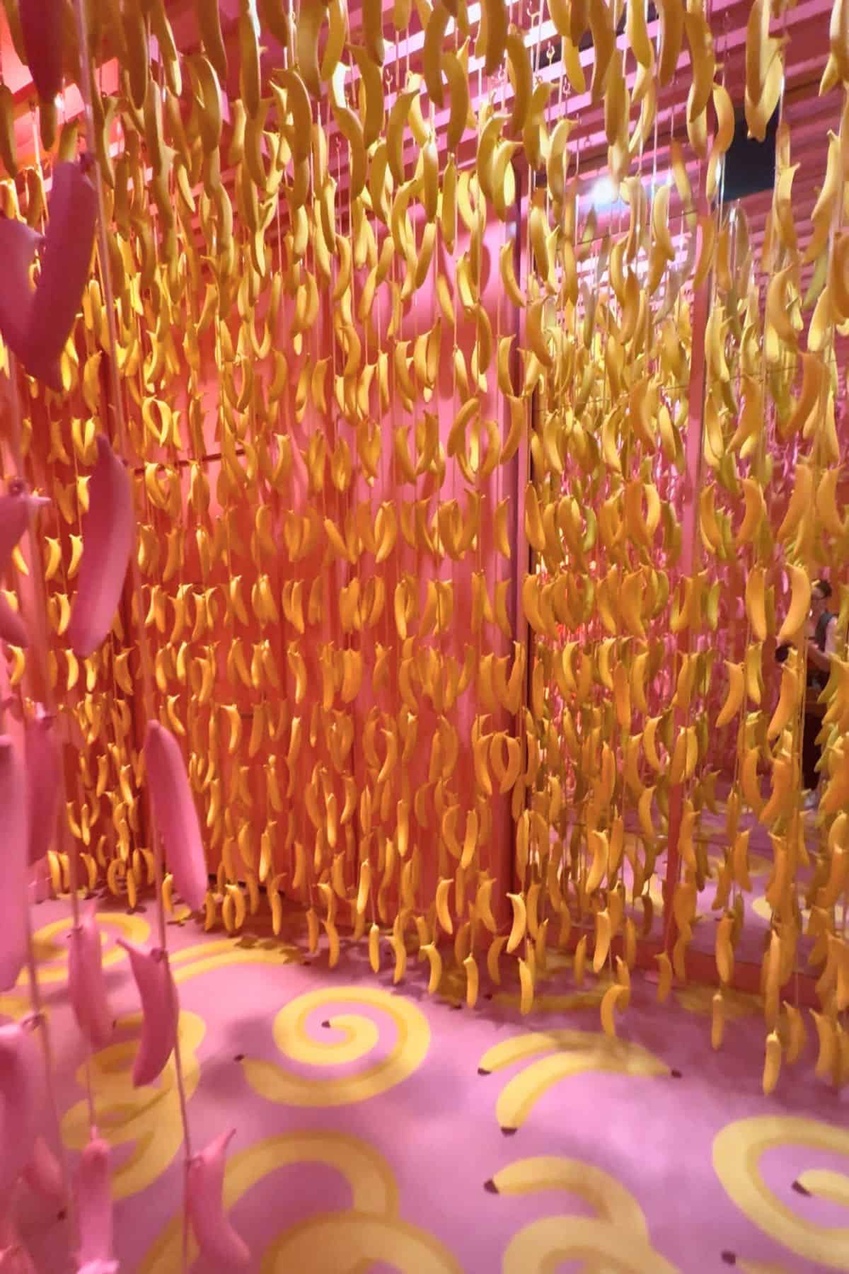 Room decorated with hanging yellow and pink banana-shaped objects, pink walls, and a floor with yellow spiral patterns.