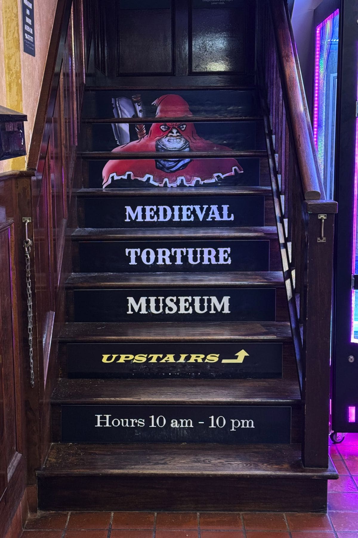 Wooden staircase with a red-hooded figure cutout and signs reading "Medieval Torture Museum Upstairs," along with hours of operation from 10 am to 10 pm.