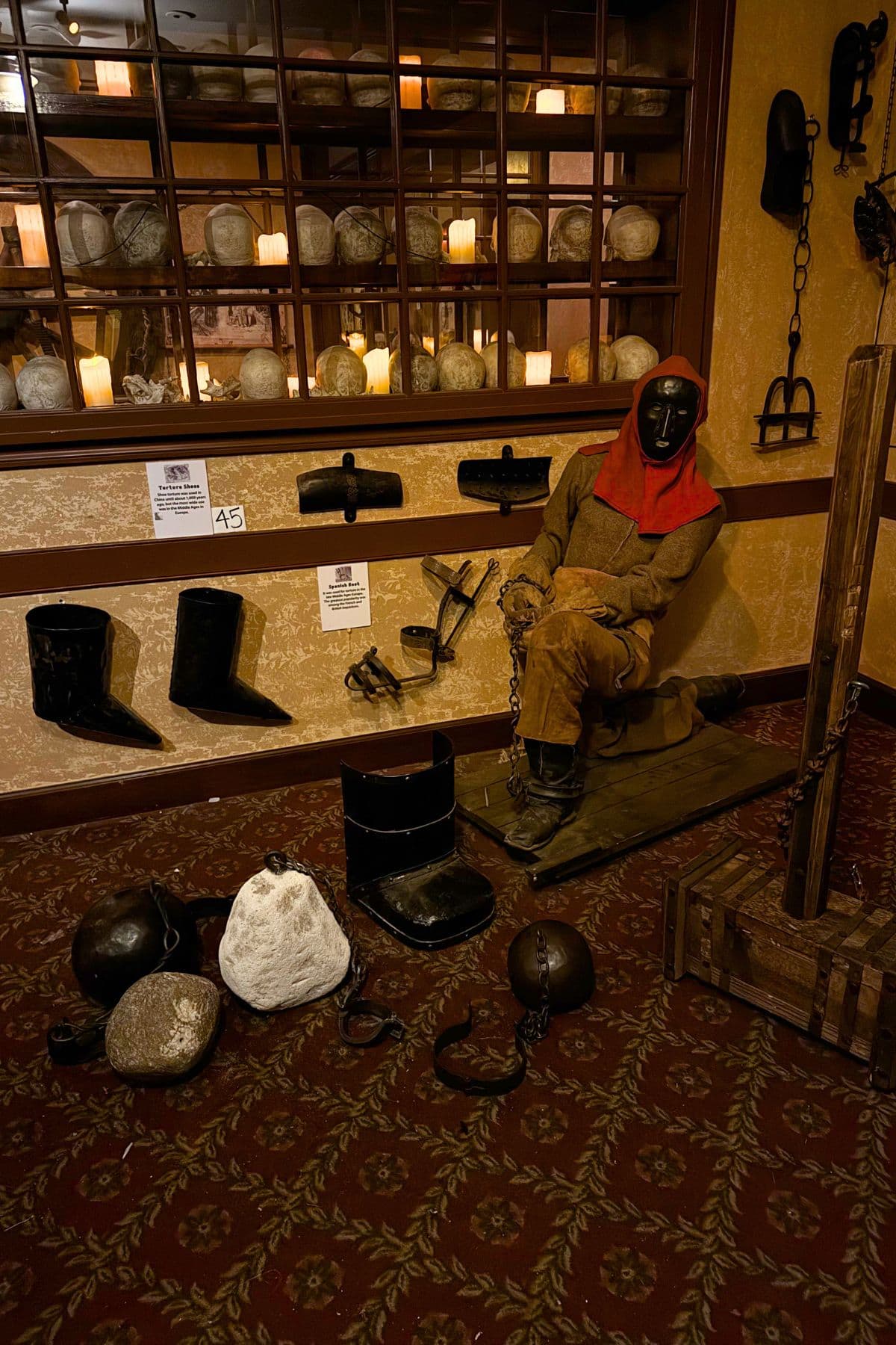 A museum display of historical torture devices features a mannequin in period clothing with a metal mask, surrounded by shackles, iron boots, and various restraints on a patterned carpet.