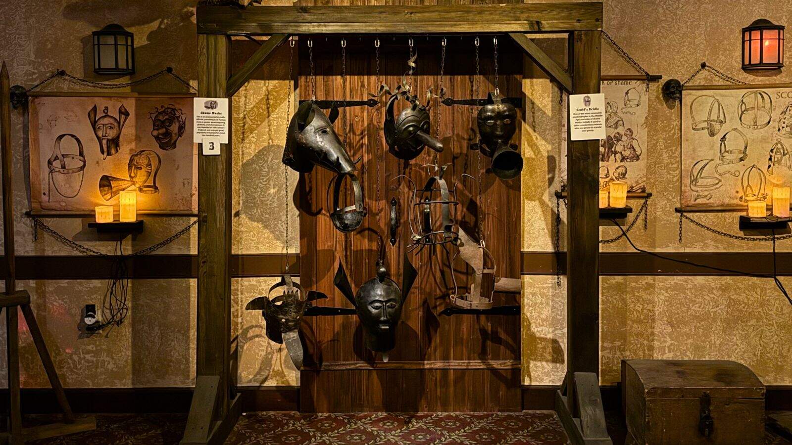A display of metal masks and headpieces hanging from a wooden frame in a dimly lit museum room, with sketches and candles on the walls in the background.