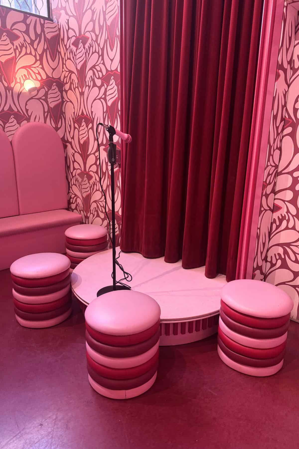 A small pink stage with a microphone stand, surrounded by round pink stools and a pink bench, set against a floral wallpaper and red velvet curtain.