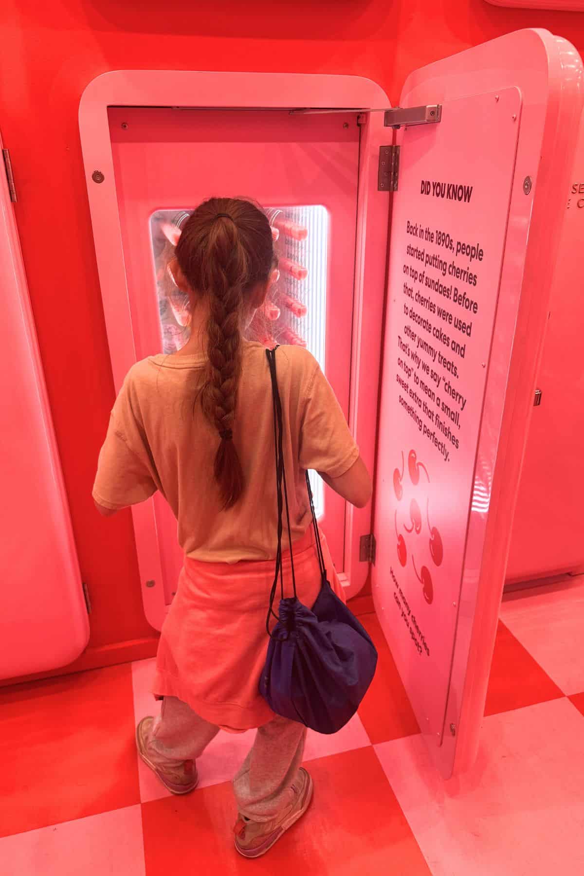 A person with long hair, wearing a peach shirt and carrying a blue bag, stands in front of an open pink refrigerator inside a brightly lit pink room.