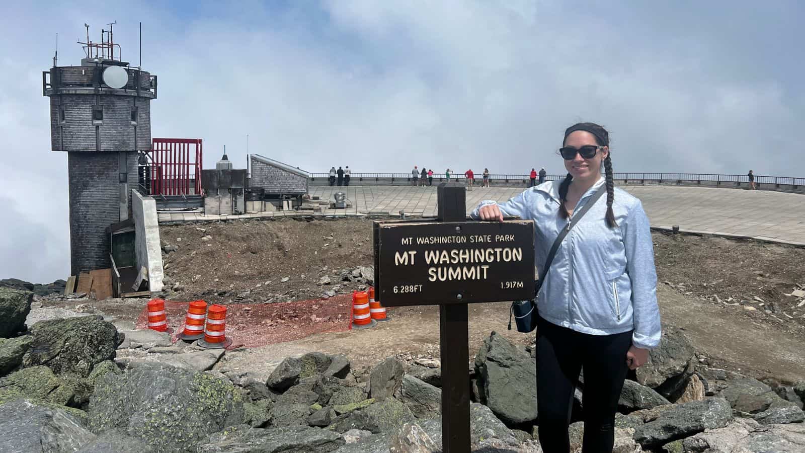 A woman stands next to the Mt. Washington Summit sign at 6,288 feet, with a weather station, rocks, and traffic cones in the background.
