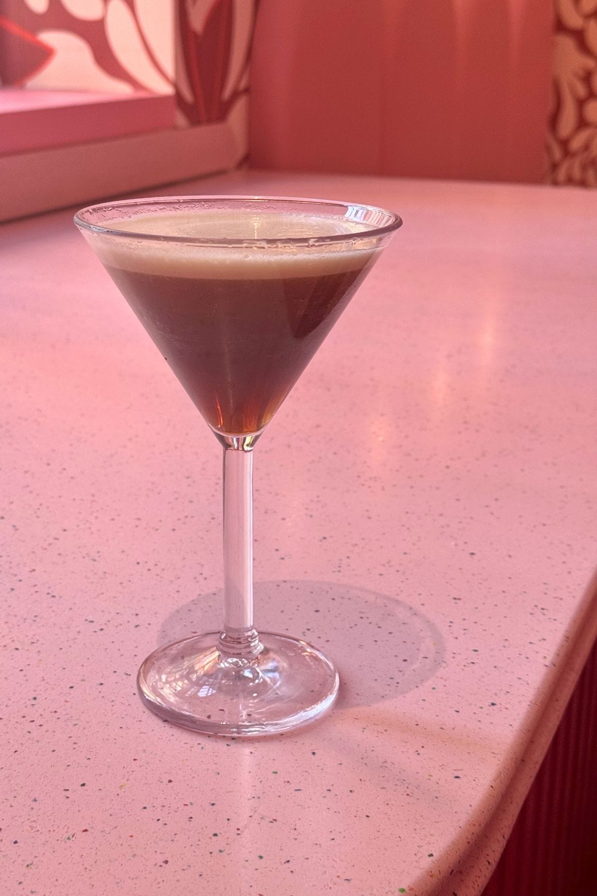 A martini glass filled with a dark cocktail topped with foam sits on a pink speckled countertop in a pink-toned room.