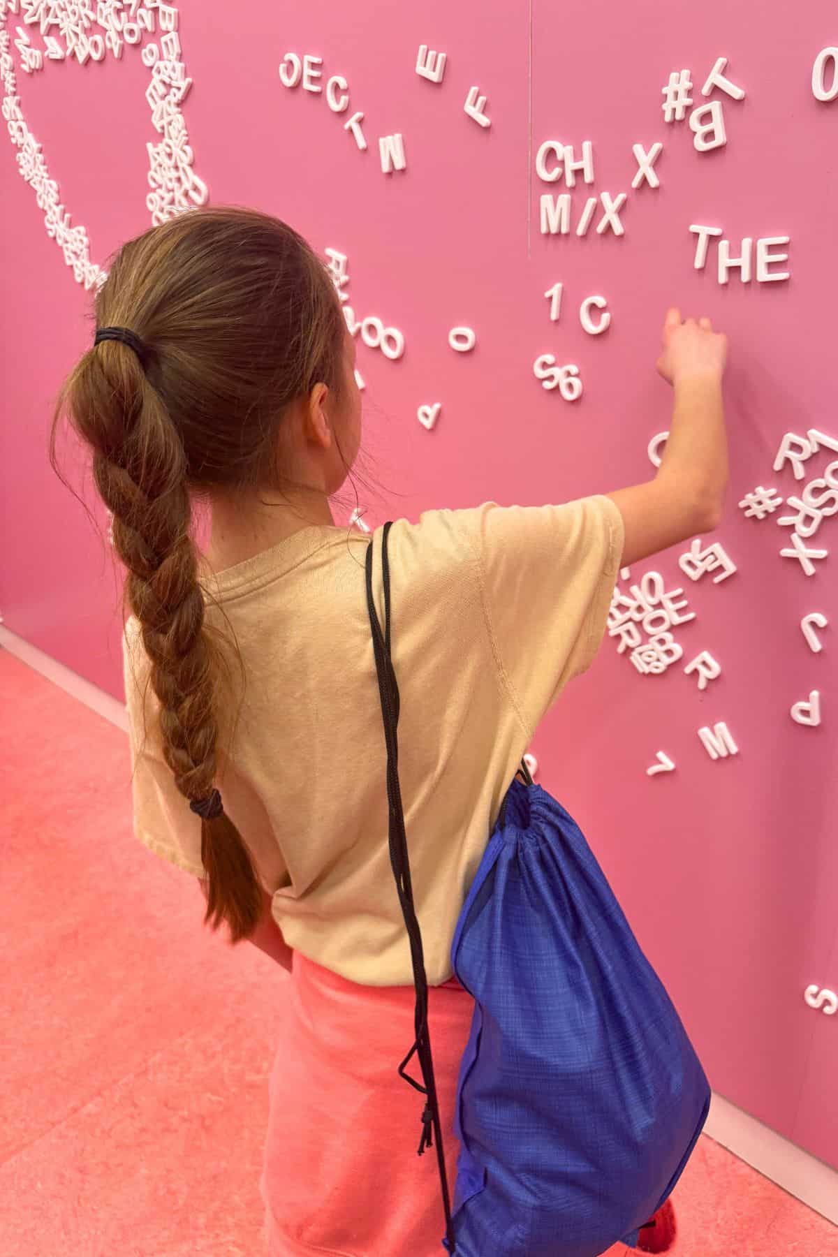 A girl with a braid and blue drawstring bag arranges white magnetic letters on a pink wall.