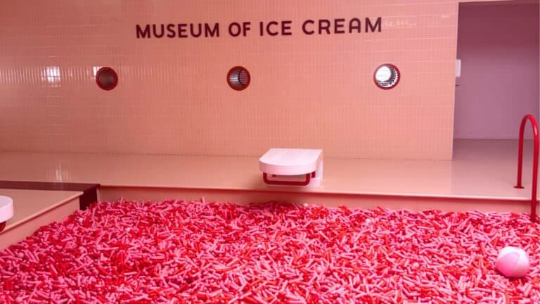 A room labeled "MUSEUM OF ICE CREAM" features a large pool filled with pink and red plastic sprinkles and minimal, modern furnishings.