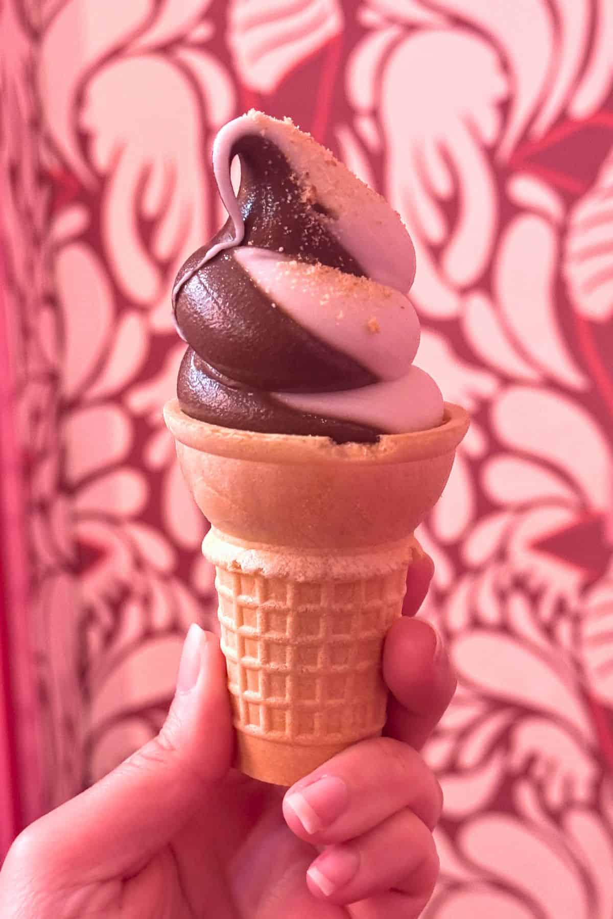 A hand holds a waffle cone with swirled chocolate and lavender soft serve ice cream against a pink and white patterned background.