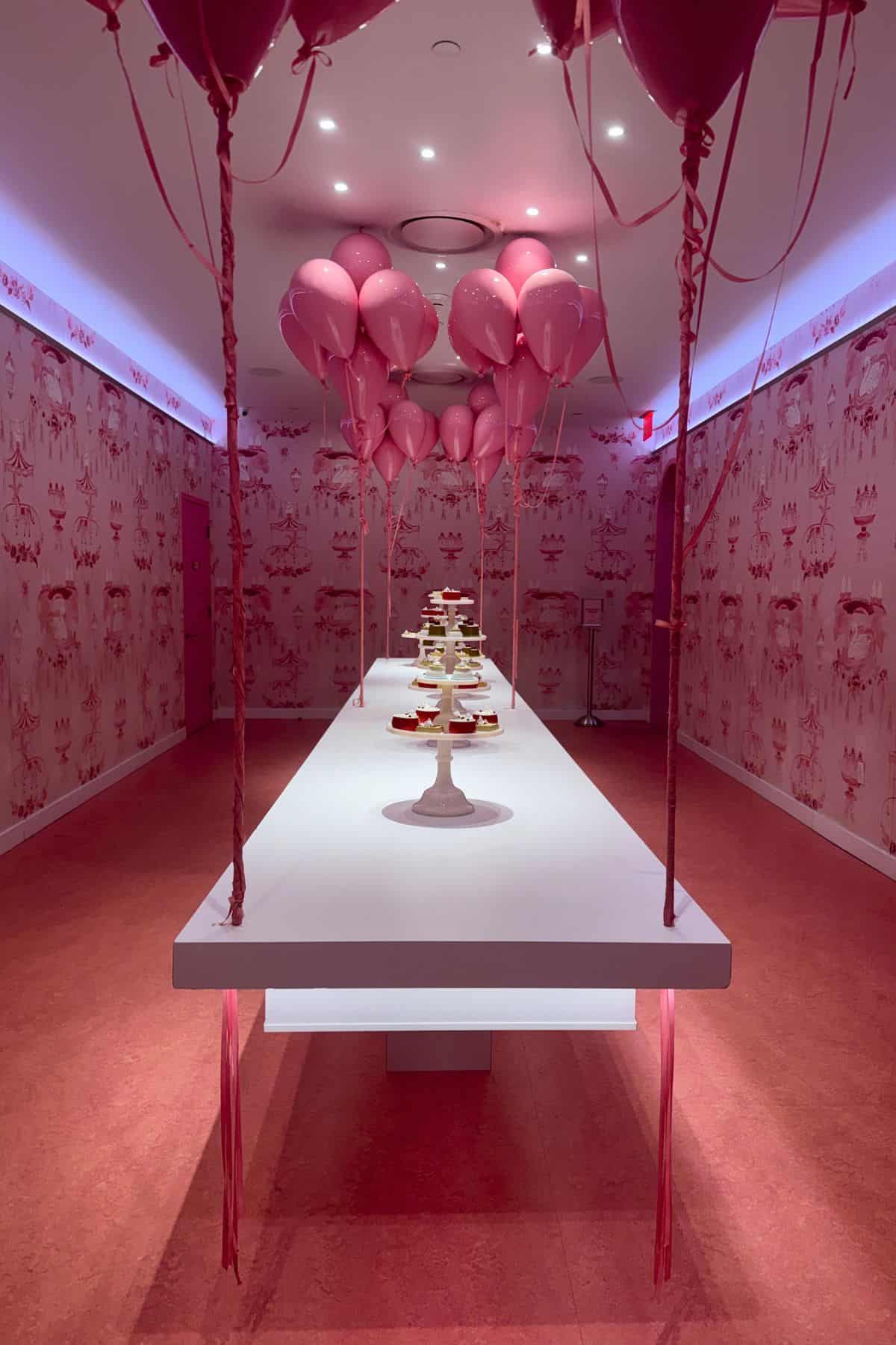 A long white table with stands of cupcakes is centered in a pink room decorated with pink balloons and patterned wallpaper.