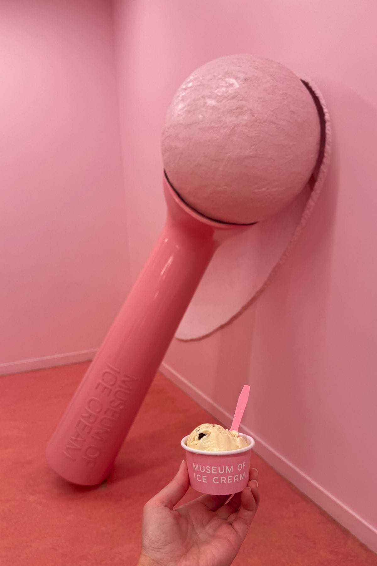 A hand holds a cup of ice cream in front of a large pink ice cream scoop sculpture at the Museum of Ice Cream, with pink walls and flooring in the background.